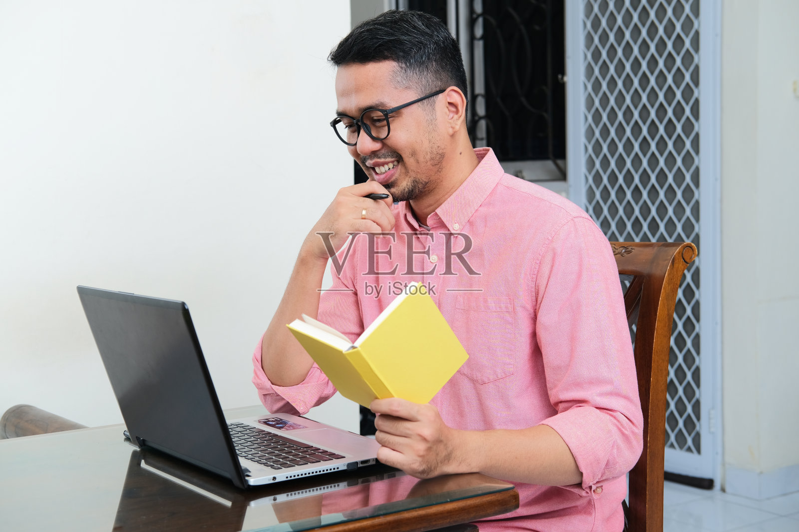 亚洲成年男子拿着一本书，看着他的笔记本电脑，脸上带着幸福的表情照片摄影图片