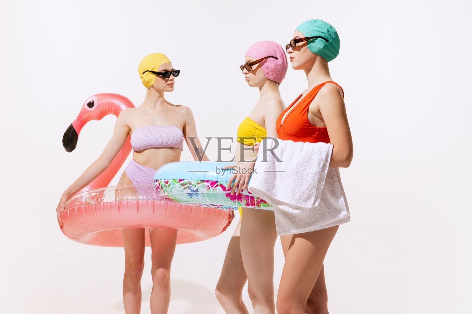 三名年轻女性在泳装，帽子和太阳镜与游泳圈孤立在灰色的工作室背景照片摄影图片