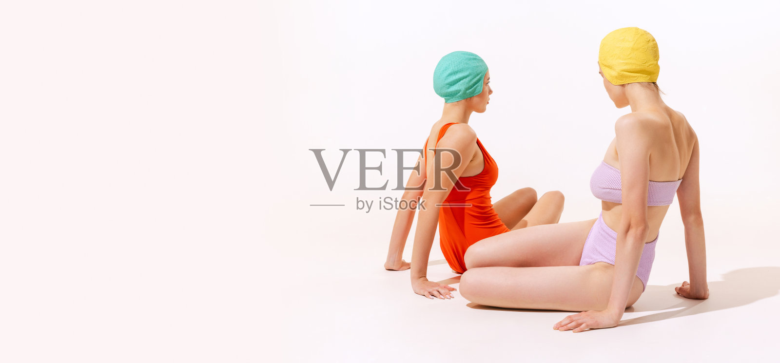 两名年轻女子在泳装和帽子坐着的肖像，在灰色的工作室背景上孤立地摆拍照片摄影图片