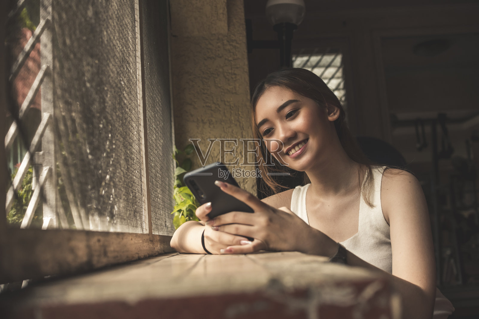 一个年轻的微笑的亚洲女人忙着在她的手机聊天，而挂在一个昏暗的房间。照片摄影图片