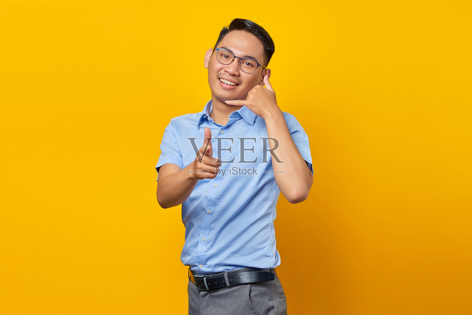 微笑的亚洲年轻男子在眼镜显示叫我的手势和手指的相机孤立的黄色背景。商人与企业家概念照片摄影图片