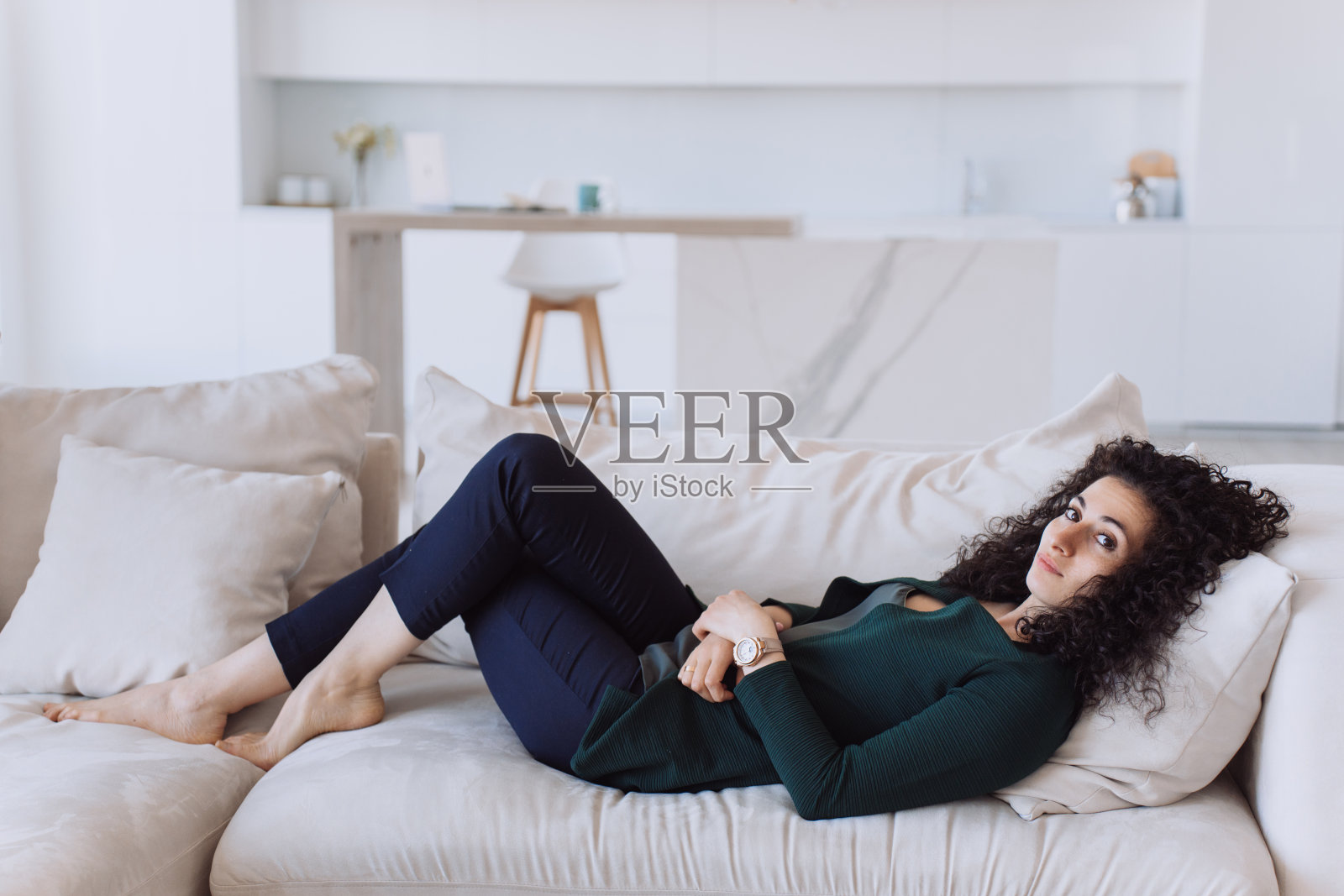 漂亮的西班牙卷发女人穿着绿色开衫，蓝色裤子躺在舒适的沙发在客厅放松家务后，看着相机腿交叉，白人黑发女人在家里休息。照片摄影图片