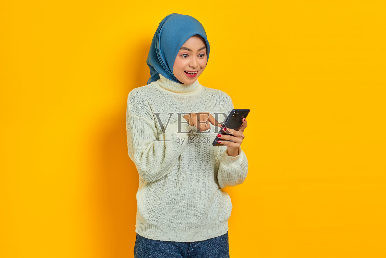 肖像开朗的年轻亚洲妇女在白色毛衣和希贾布使用手机，指着屏幕手机孤立的黄色背景。人们伊斯兰教宗教观念照片摄影图片