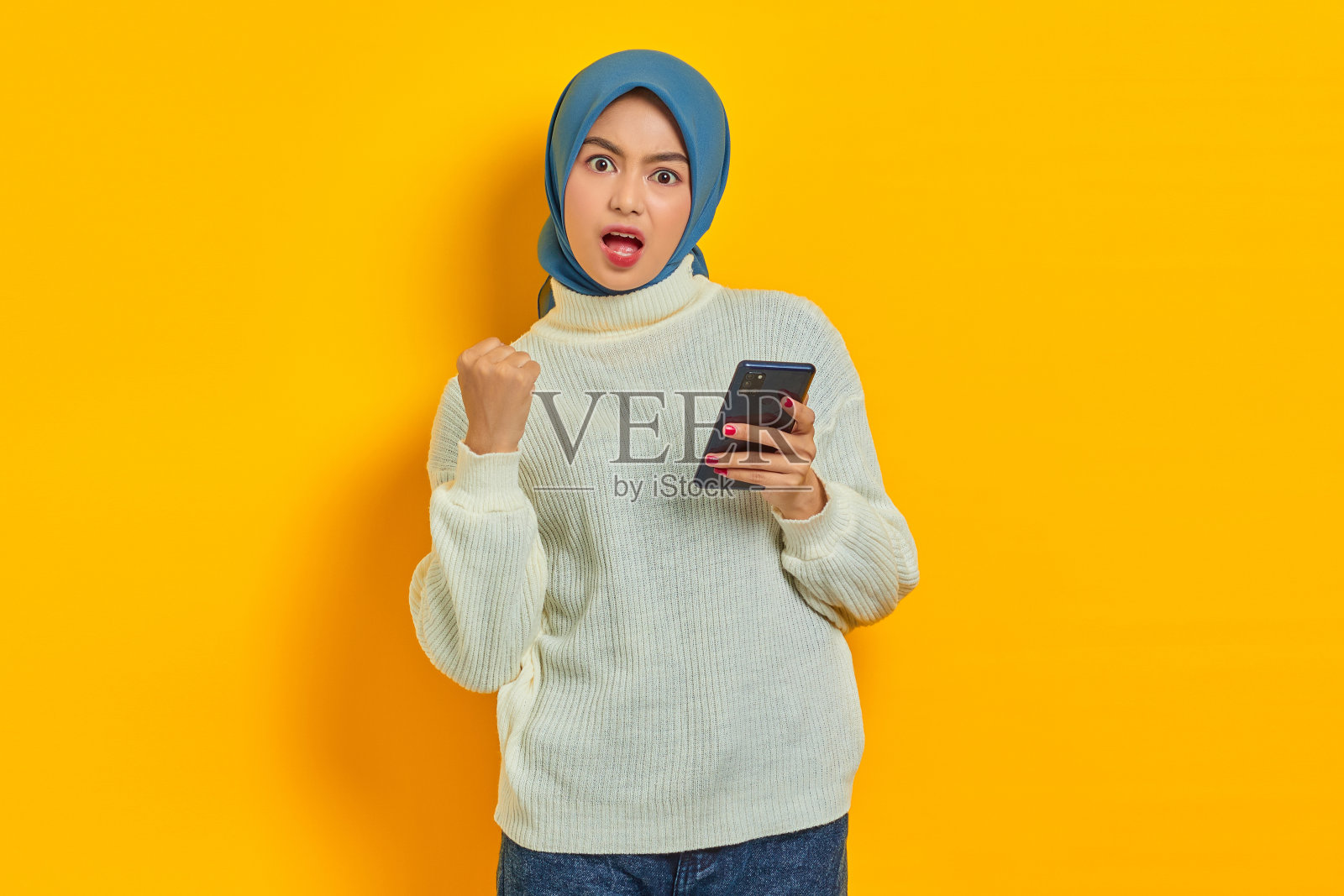 震惊的年轻亚洲女性在白色毛衣和头巾使用手机，做赢家的手势握拳说yes孤立在黄色背景照片摄影图片