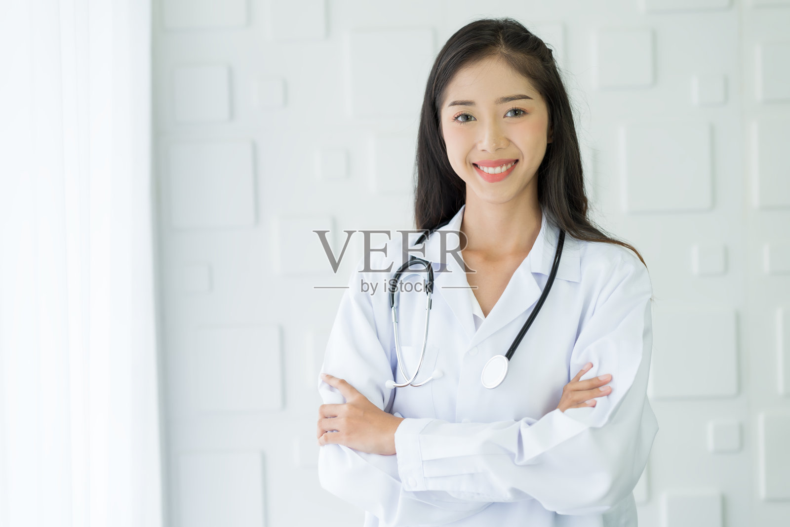 快乐的亚洲护士穿着带听诊器的制服，双臂交叉站在医院里。照片摄影图片