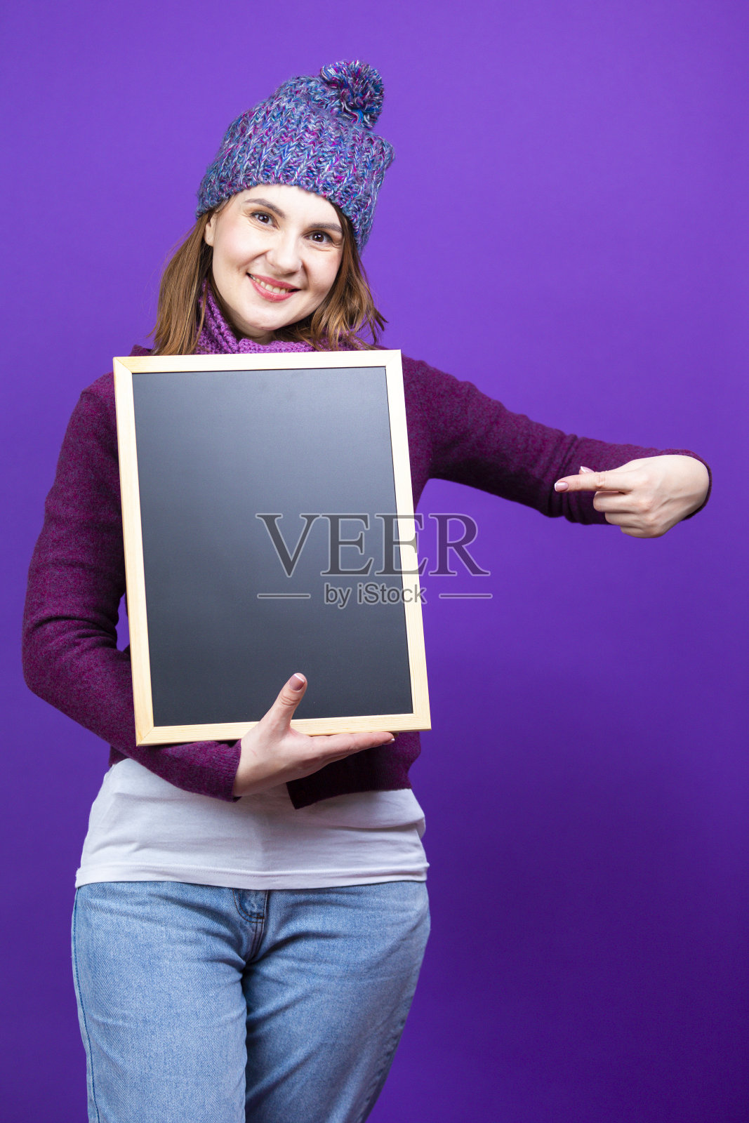 现代生活方式的概念。积极的高加索女性在温暖的针织冬季帽子和紫色围巾指向黑色木制黑板上时髦的紫色背景。照片摄影图片