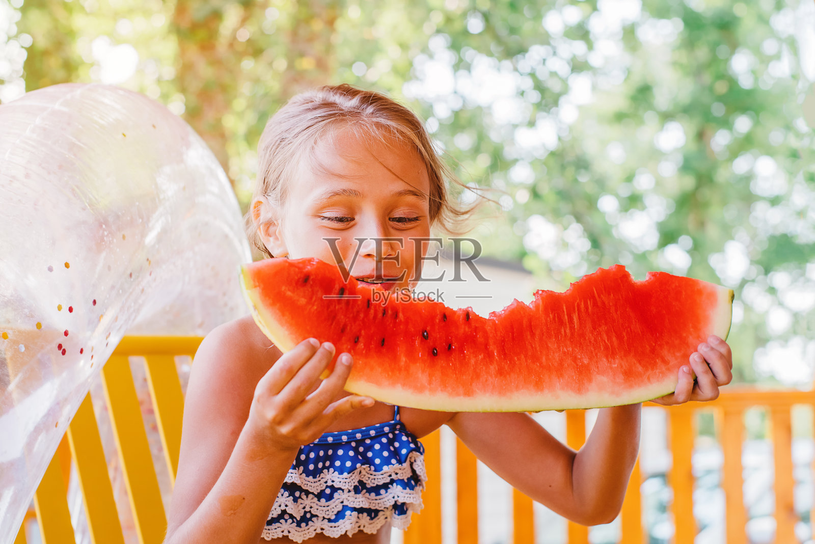夏日露台上的女孩笑着吃了一大块红色的西瓜。自然的食物。女孩在夏天吃一个成熟多汁的西瓜。可爱的金发女孩在户外吃一片西瓜。照片摄影图片