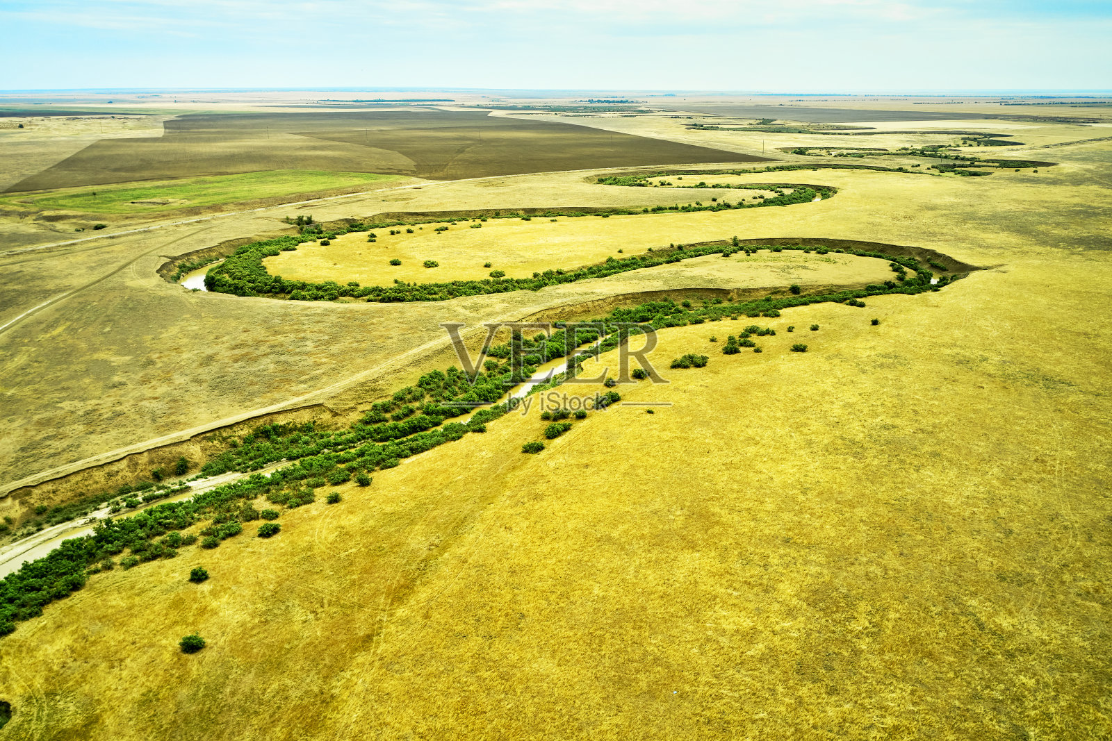 一条弯弯曲曲的泥泞河流和杂草丛生的绿色河岸，在阳光炙烤的草原乡村景观照片摄影图片