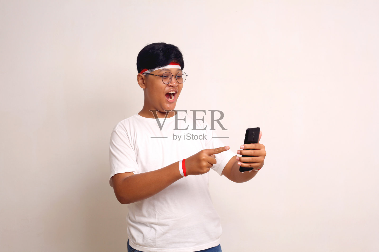 惊讶的亚洲男孩站着，拿着手机指着它。孤立在白色照片摄影图片