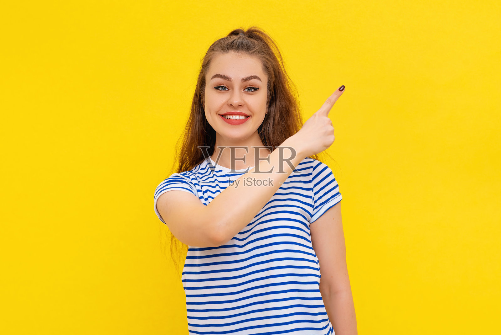 开朗的年轻黑发女子穿着休闲的白蓝条纹t恤微笑着指着右边，展示广告。室内工作室拍摄的黄色背景照片摄影图片