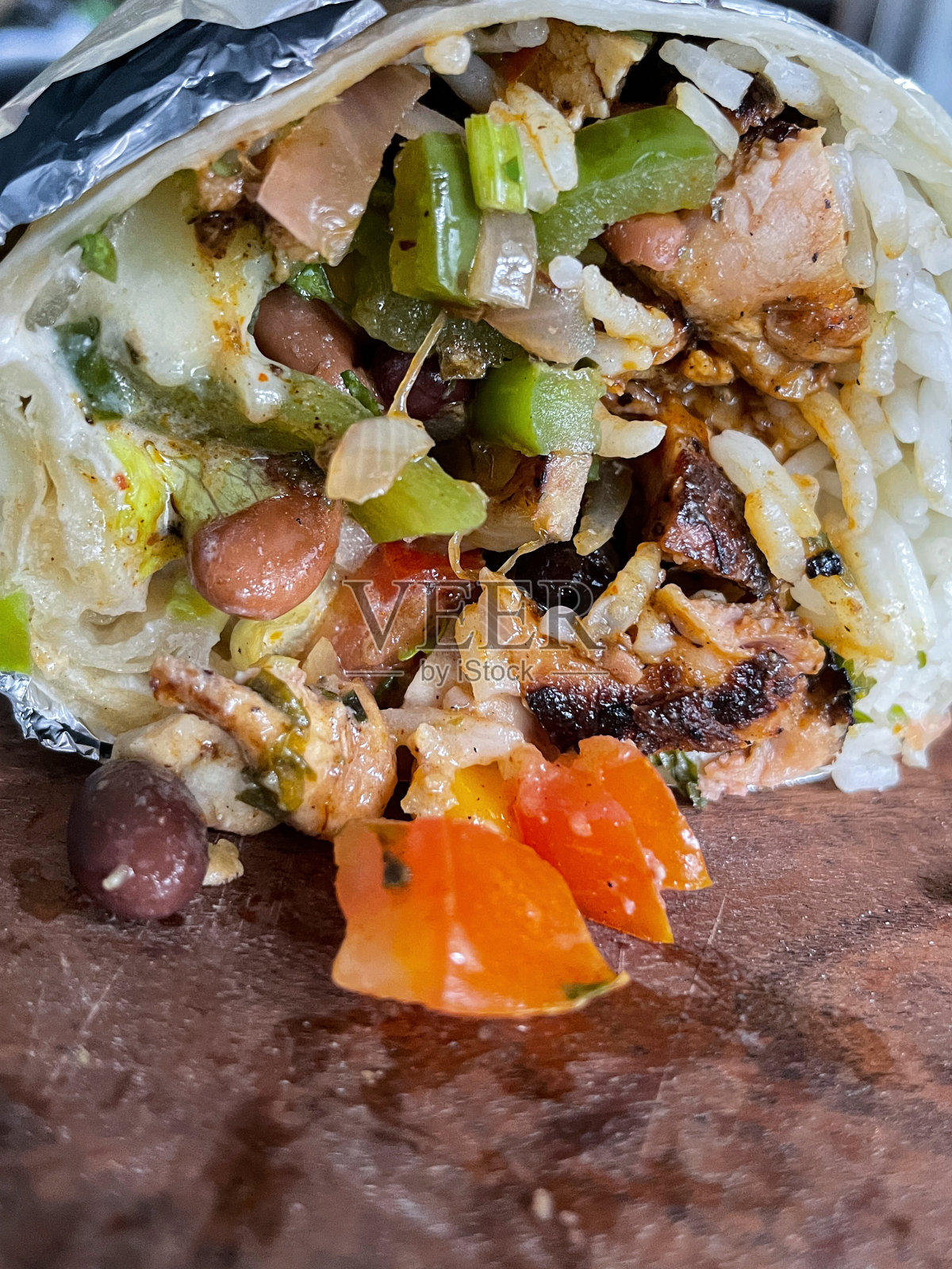 特写横截面图像的卷，自制的，软小麦玉米饼面饼卷饼一半包裹在锡纸，大米，鸡肉，甜玉米，黑色和斑豆，黄瓜，红青椒馅，在木制砧板上，重点放在前景照片摄影图片