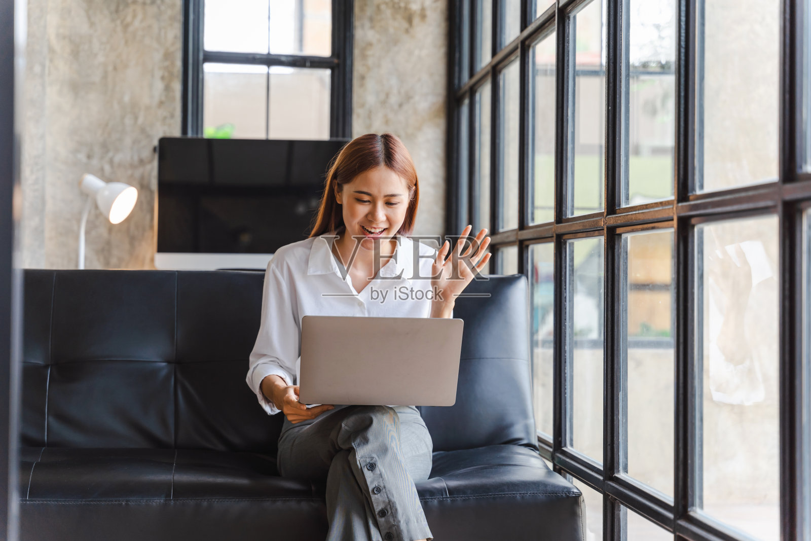 年轻的亚洲女性坐在沙发上，使用笔记本电脑，在互联网和社交媒体上向朋友挥手致意。照片摄影图片