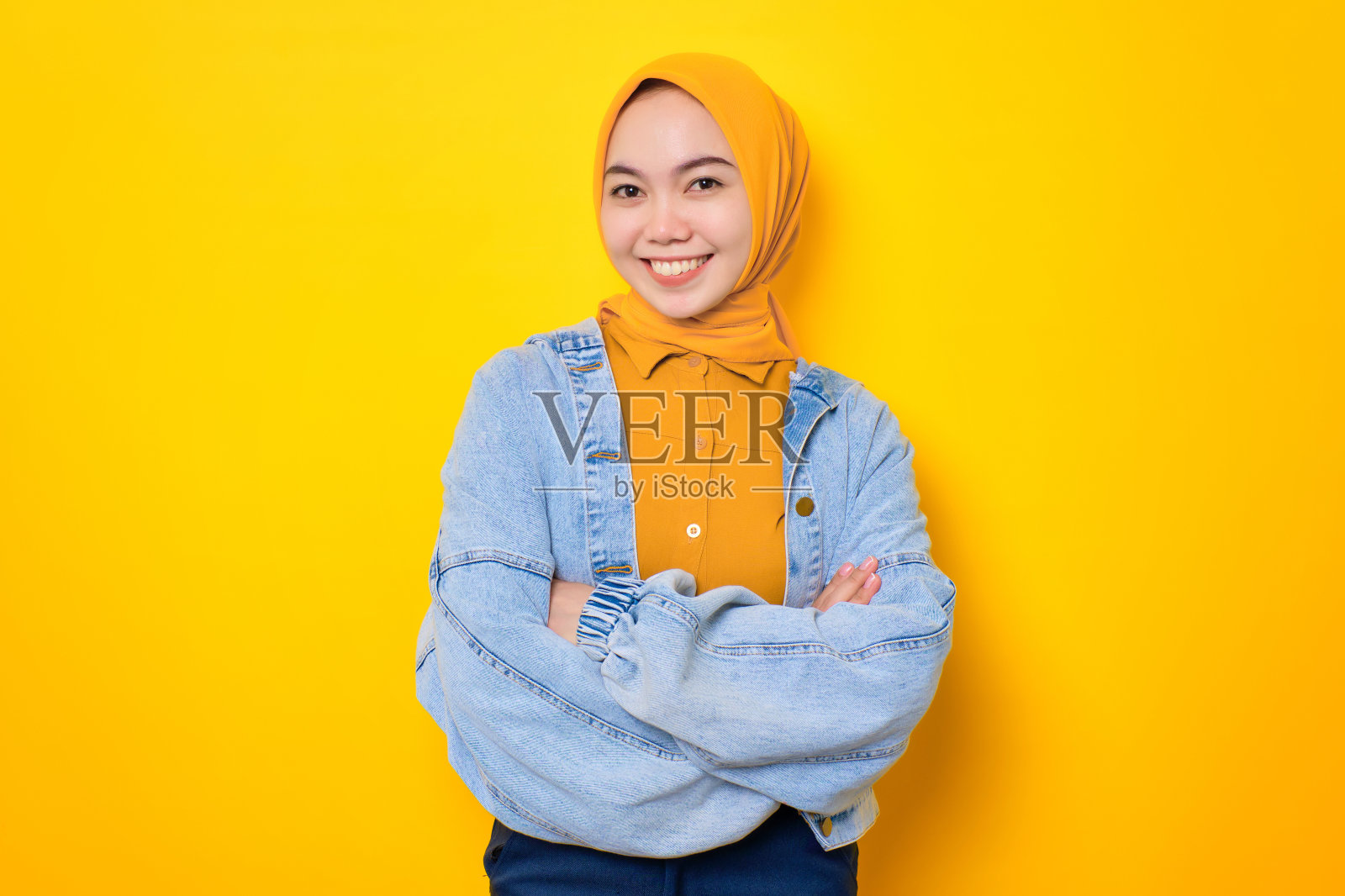 快乐的年轻亚洲女人在牛仔裤夹克交叉双臂，看起来自信孤立在黄色背景照片摄影图片