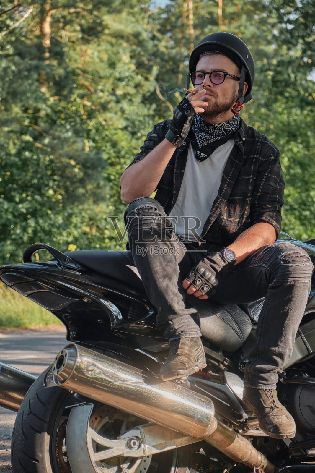 年轻的男性摩托车手戴着头盔独自骑着摩托车旅行，停在森林道路的一边吸烟照片摄影图片