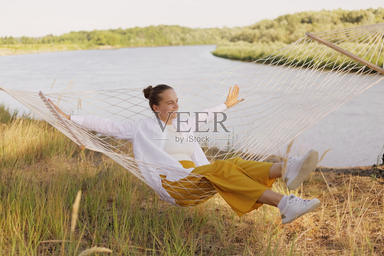 户外拍摄的微笑满意的女性穿着白衬衫坐在吊床上的河岸边，摊开双手，看向远处的美丽的自然。照片摄影图片