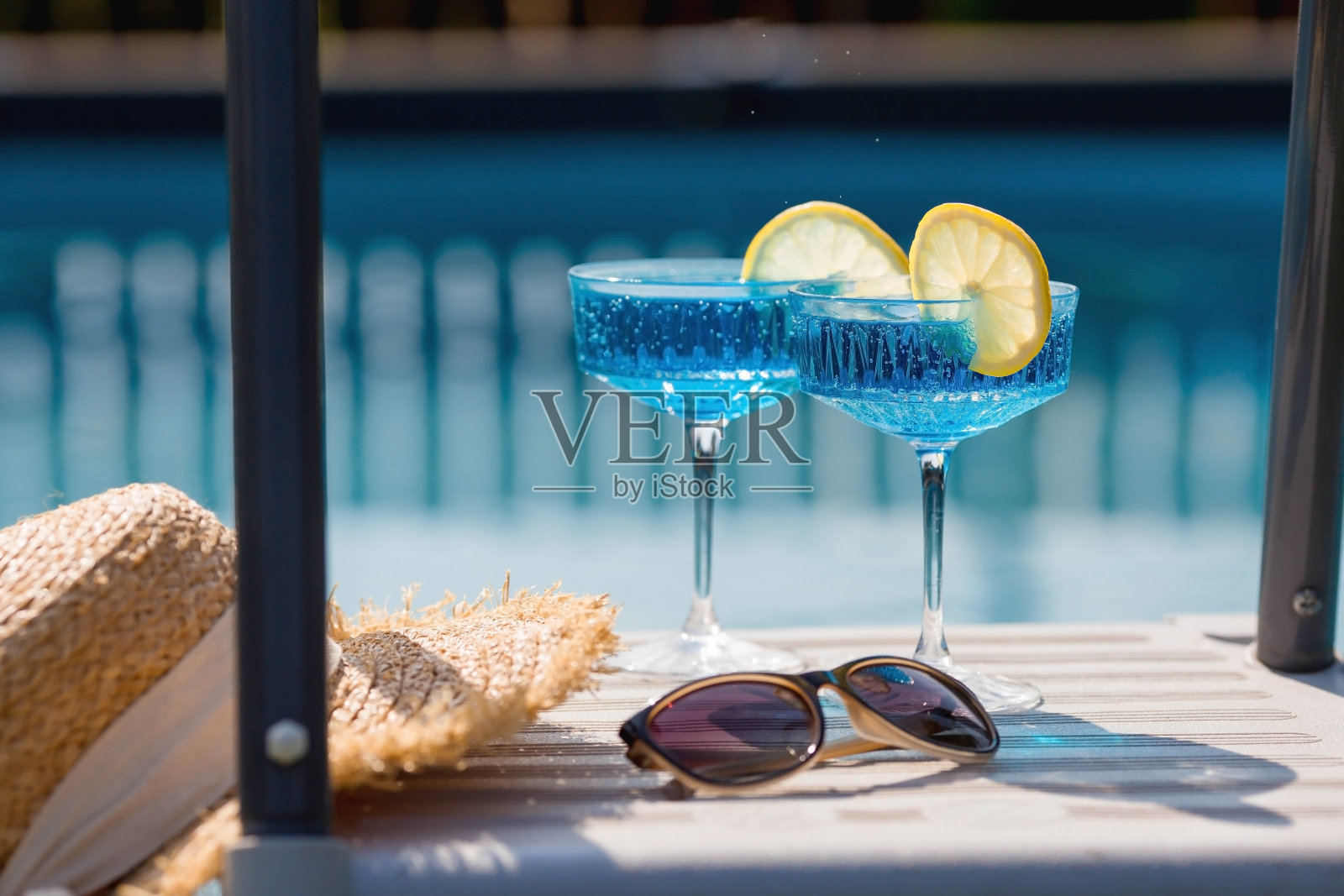 一杯美味的蓝色鸡尾酒“蓝色泻湖”和黄色的柠檬放在泳池的背景上。酒精鸡尾酒多汁的水果蓝色与库拉索利口酒，冰块和一片柠檬。照片摄影图片