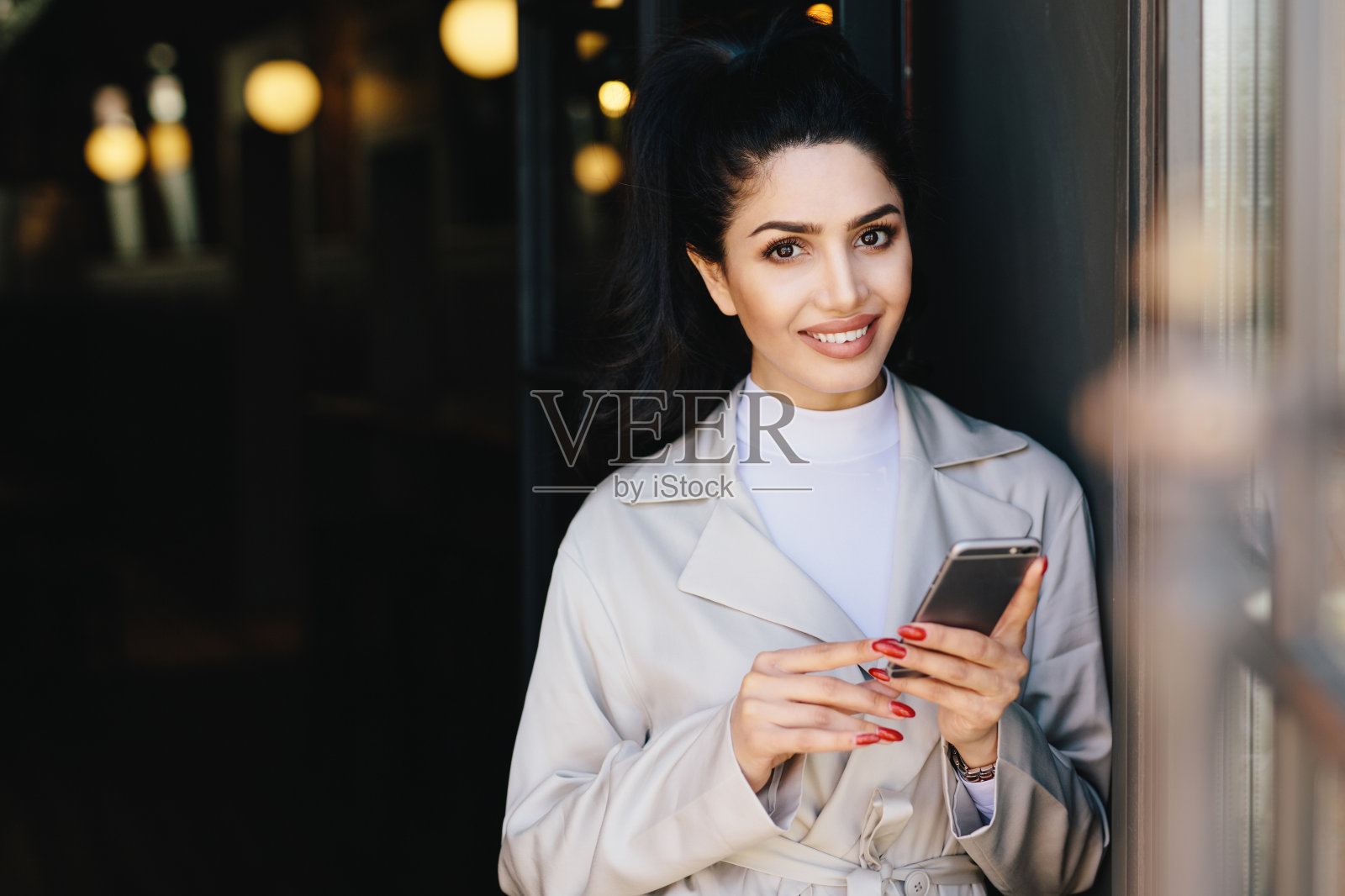 性感的黑发女士，迷人的黑眼睛和丰满的嘴唇，红色的指甲拿着手机，与她的朋友交流，微笑，看着相机。时尚的概念照片摄影图片