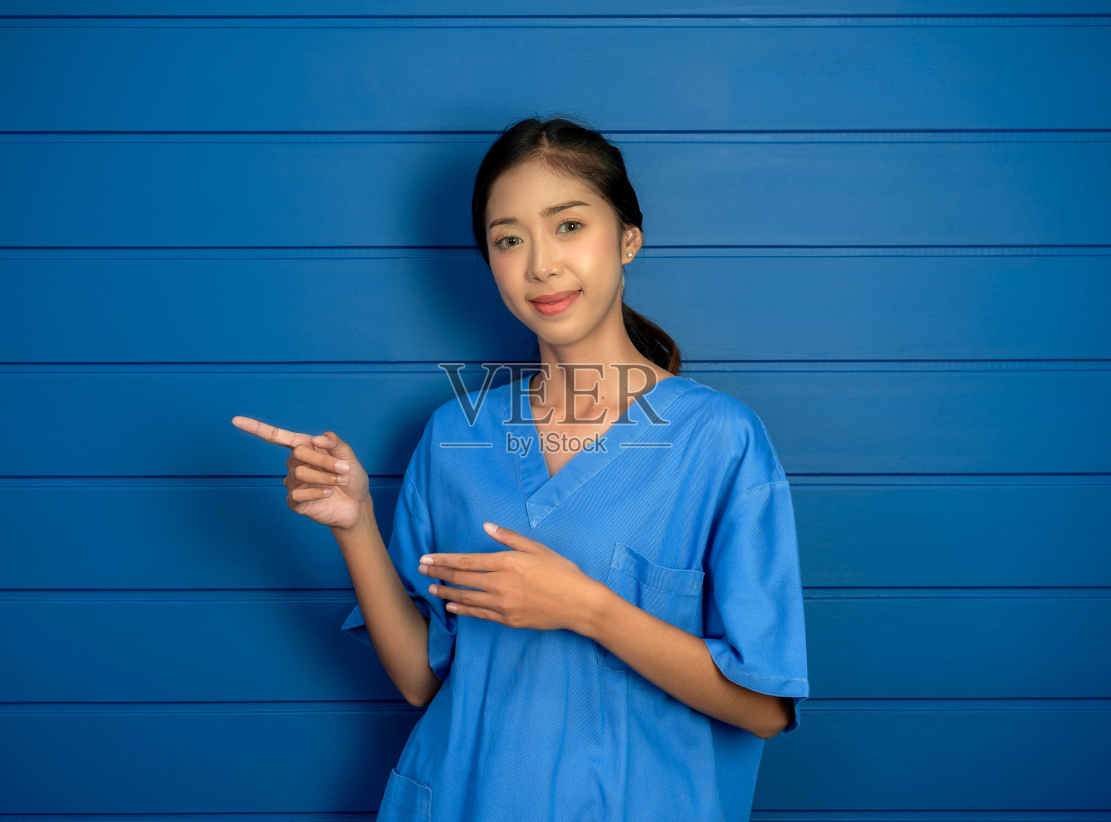 穿着蓝色手术服的友好、微笑、自信的亚裔女医生或护士手指向左的肖像，显示在蓝色的木制墙壁背景上，宣传保健和医疗。照片摄影图片