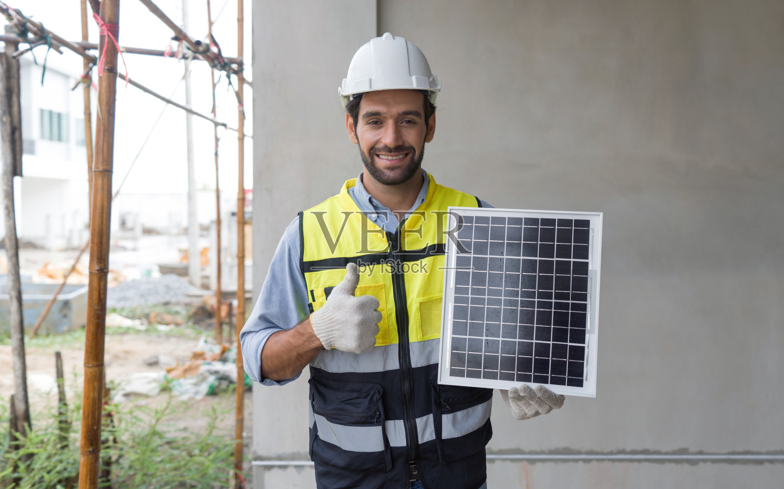 年轻的工程师戴着安全帽、安全背心和手套，拿着太阳能电池板，微笑着举起大拇指，展示一种能产生直流电的能源。照片摄影图片