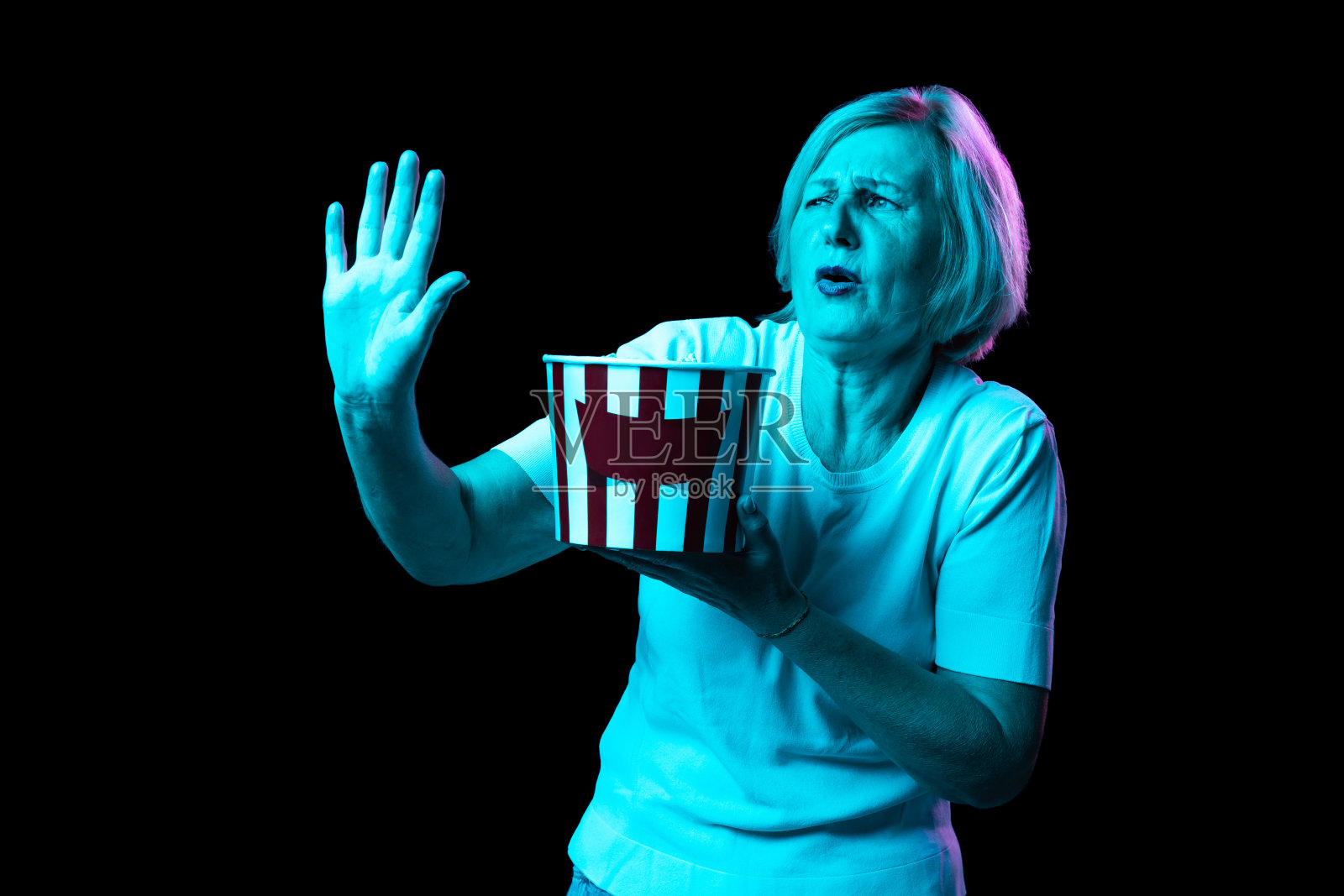 一个兴奋的高级妇女，现代祖母拿着一桶爆米花孤立在黑暗的背景霓虹灯。情绪的概念，面部表情，渴望照片摄影图片