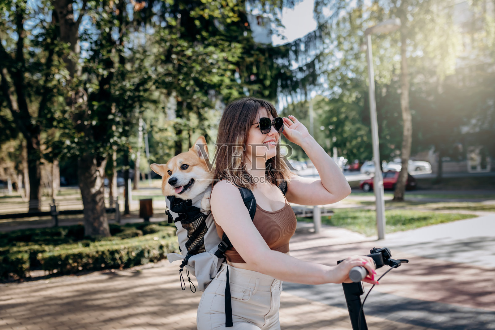 快乐微笑的女游客在城市公园里骑着她的电动滑板车，她的背包里装着一只威尔士柯基犬照片摄影图片