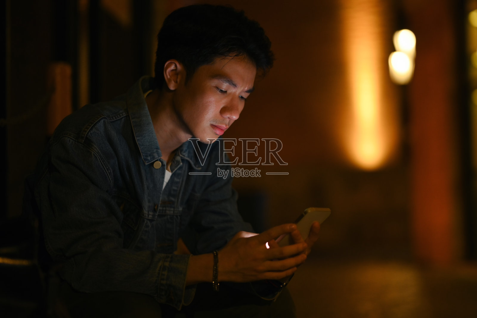 年轻的潮男坐在夜晚的城市街道上，用手机和朋友在社交网络上聊天。照片摄影图片