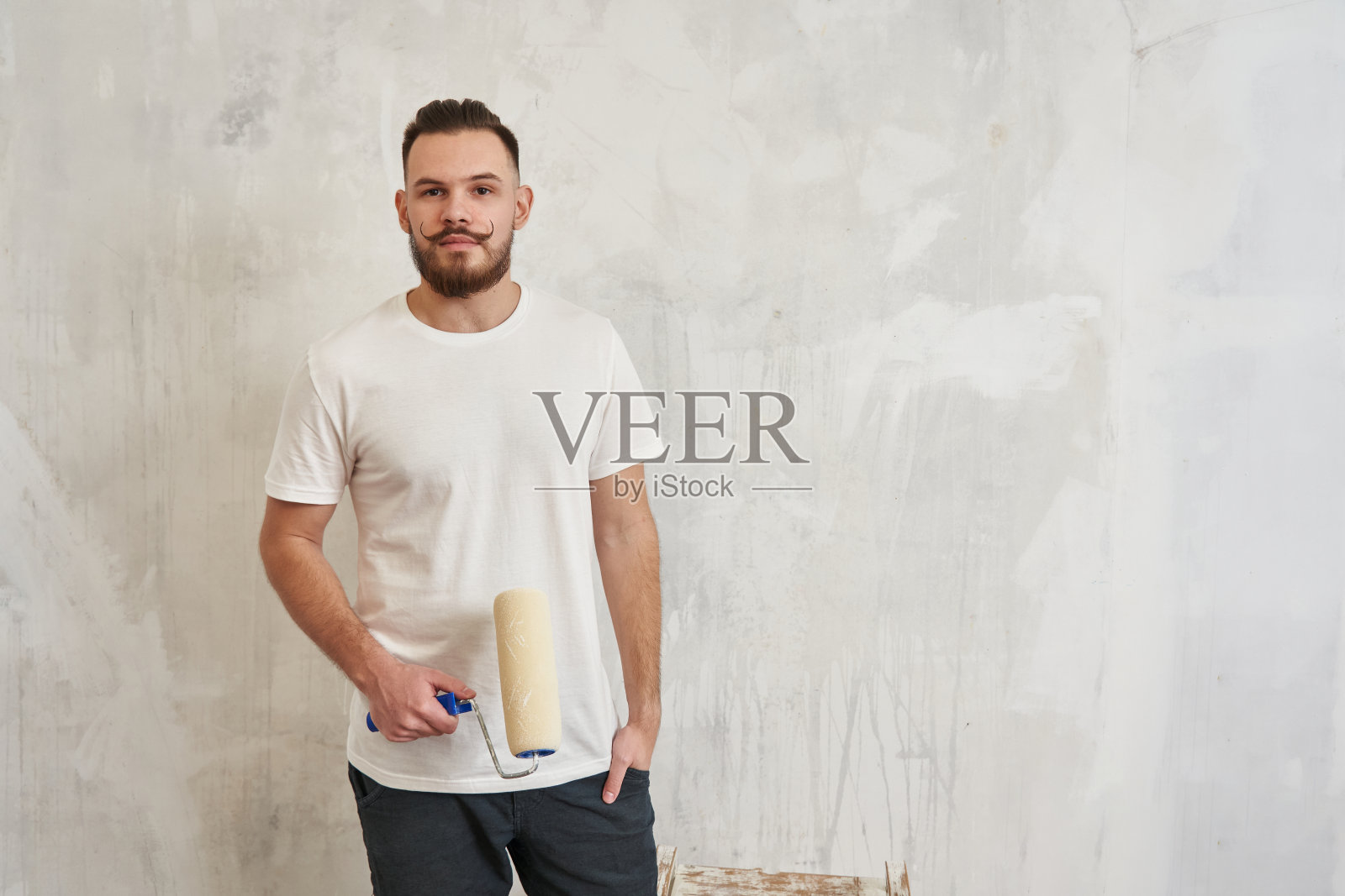 一个年轻人拿着一个涂色滚筒，背景是一堵白色的墙。维修、施工、刷墙照片摄影图片