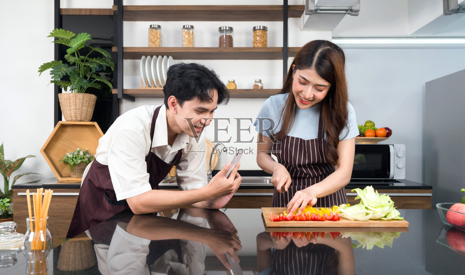 亚洲夫妇在厨房共度时光。身穿围裙的年轻女子在做沙拉，而他的男朋友在为社交博客录制视频。现代生活方式人们的关系和活动照片摄影图片