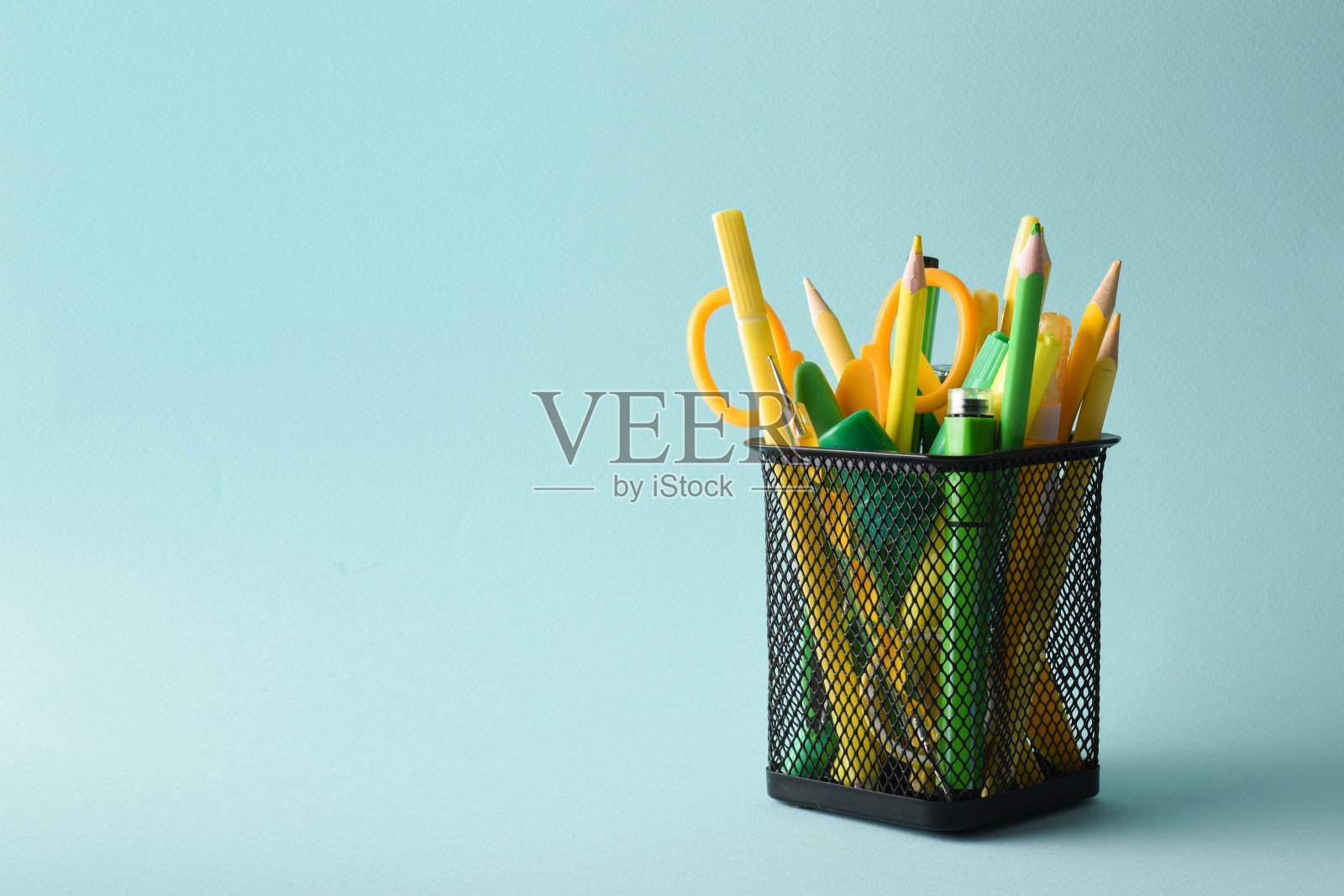 一个黑色的金属罐子，里面有黄色和绿色的钢笔、颜料和铅笔，背景是浅蓝色的。学习用品。复制的空间。垂直摄影照片摄影图片