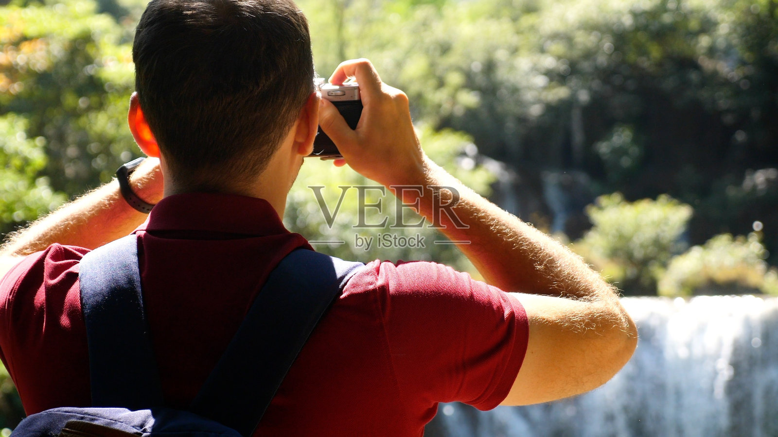 漫游癖、旅游概念。快乐的男性游客与背包旅行。年轻的旅行者使照片美丽的异国情调的瀑布和热带自然在复古风格的相机。特写,后视图照片摄影图片