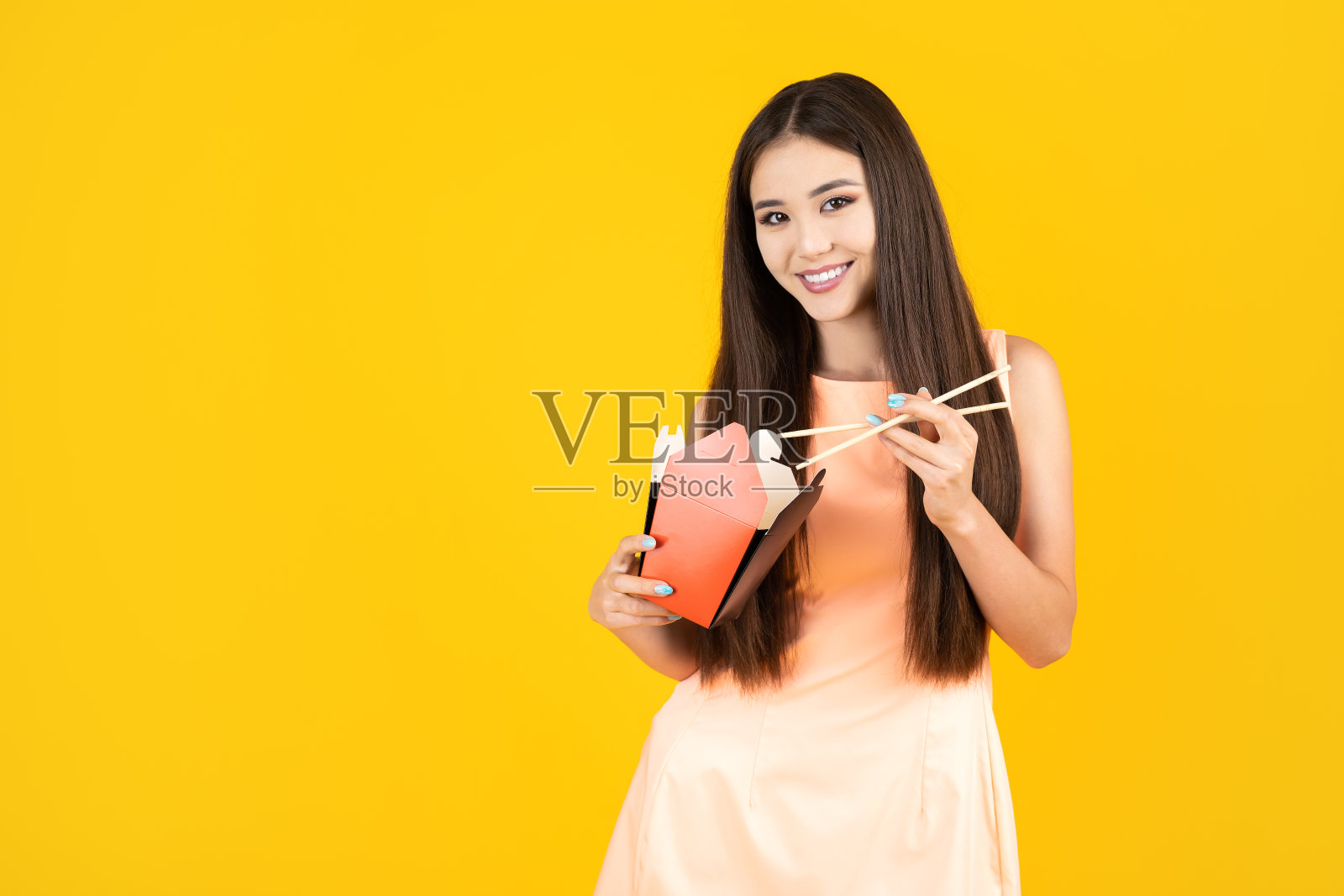 年轻迷人的亚洲女人吃亚洲炒锅食物从纸炒锅盒使用筷子在黄色的背景与拷贝空间。照片摄影图片