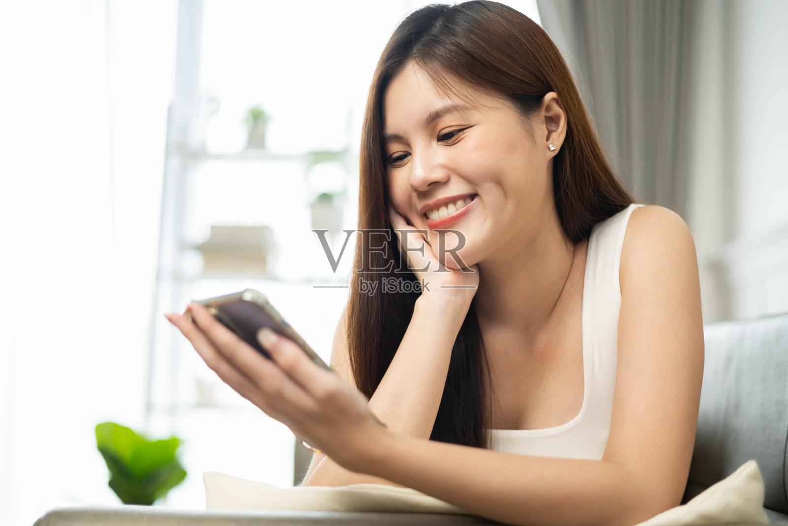 快乐而开朗的女人坐在沙发上用电话。亚洲年轻女性看着手机庆祝与好消息或折扣券网上购物在家里。照片摄影图片