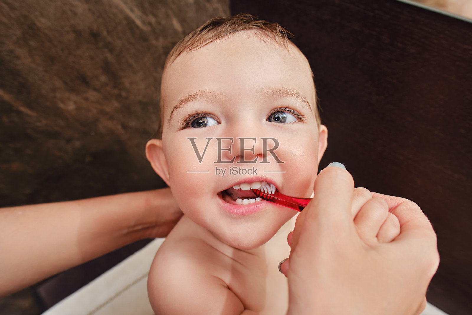 妈妈刷牙给快乐蹒跚学步的小男孩。妈妈给一个微笑的孩子刷牙。一岁的孩子照片摄影图片