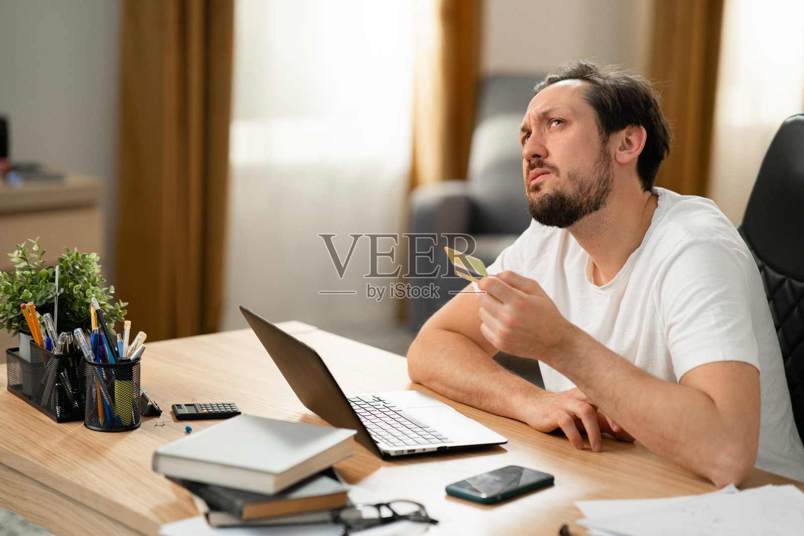 黑发留胡子的女人坐在桌子前的笔记本电脑前。他拿着银行卡，想在网上下单。他会考虑点什么更好。桌子上有电话计算器本照片摄影图片