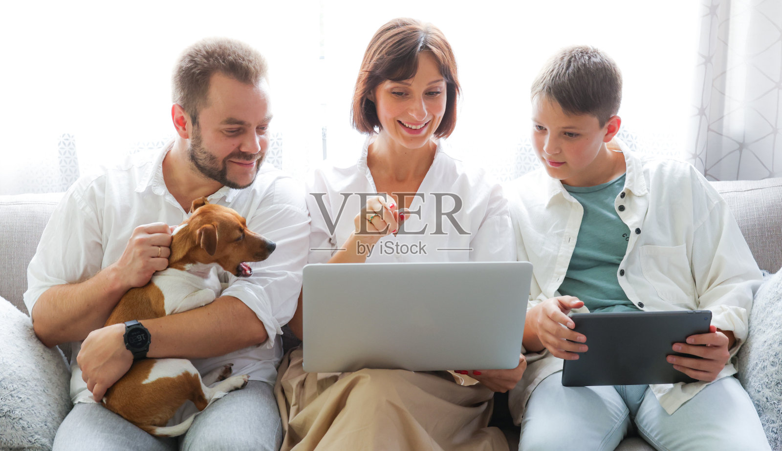 家里的沙发上放着平板电脑和笔记本电脑。妈妈，爸爸和儿子一起在家使用笔记本电脑平板电脑。网上购物、网上冲浪、视频叫网络游戏的概念。照片摄影图片