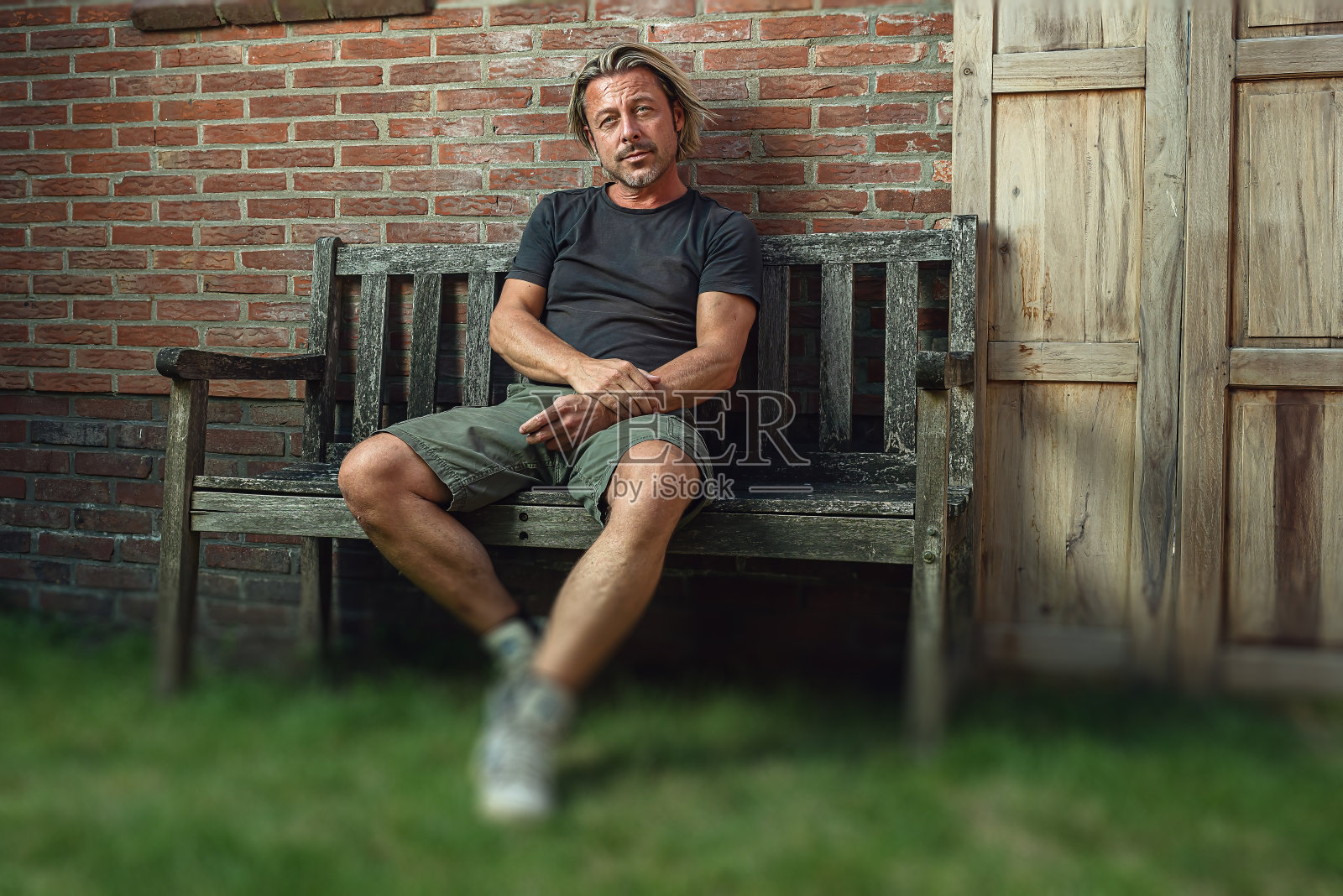 穿着黑色t恤、绿色短裤和白色运动鞋的金发男人坐在后院的长凳上。照片摄影图片