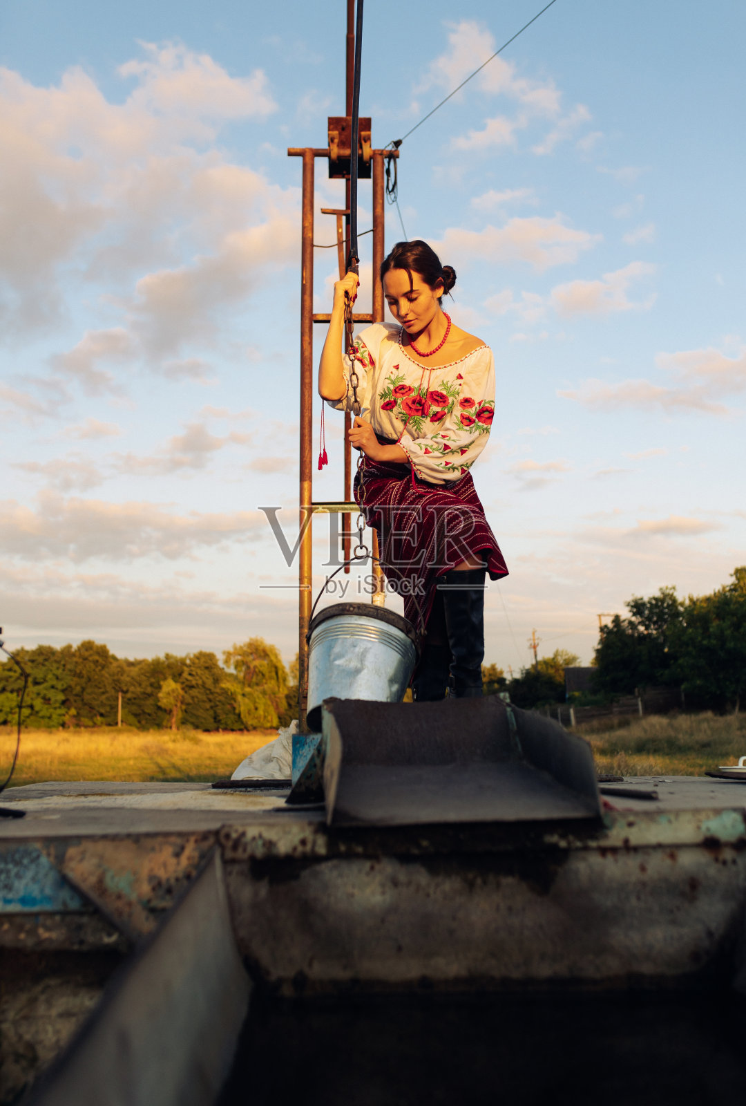 年轻的乌克兰妇女穿着传统民族服装站在草地上从井里打水。照片摄影图片