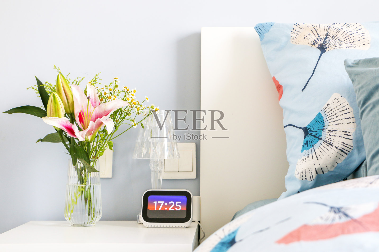 美丽的花束与盛开的粉色百合在玻璃花瓶在床头柜装饰优雅和现代的卧室淡蓝色调色板在日落，下午17:25数字时钟照片摄影图片