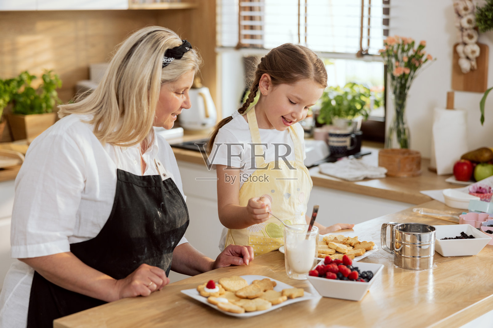 快乐的奶奶和孙子在厨房里烤饼干，站在桌子旁用甜奶油装饰饼干。为母亲准备早餐的妇女。年轻女孩玩得很开心照片摄影图片