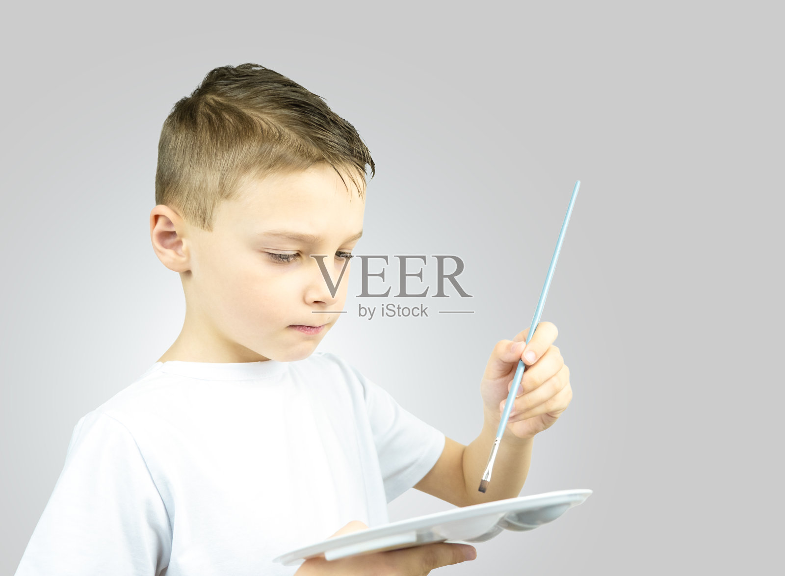 孩子的肖像谁持有画笔和调色板在他的手。穿着灰色背景白t恤的男孩照片摄影图片