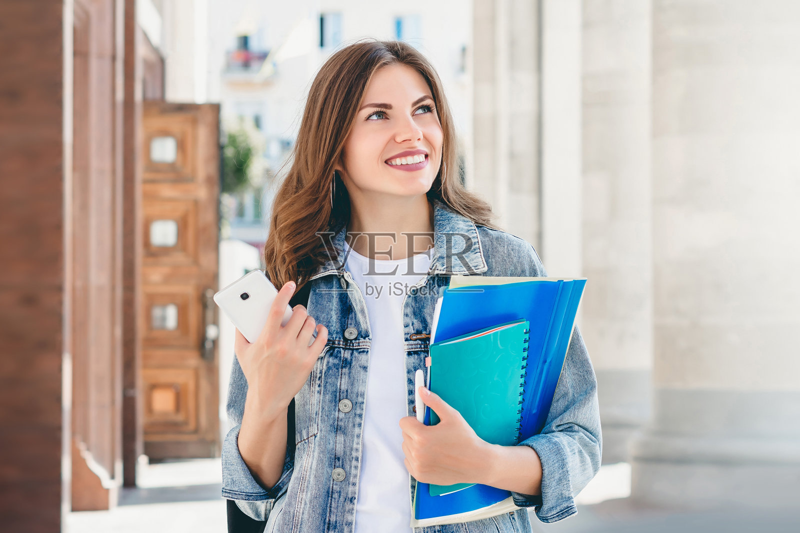 年轻的女学生对着大学微笑。可爱的女学生拿着文件夹，笔记本和手机照片摄影图片