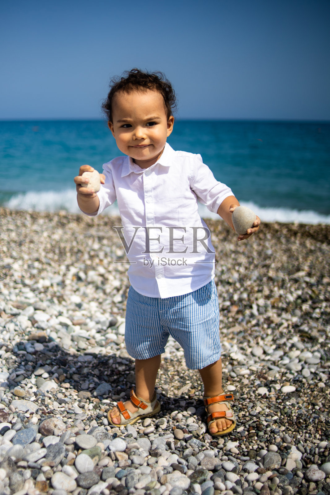 毛毛蹒跚学步的小男孩在蓝色大海附近的卵石海滩上玩耍照片摄影图片