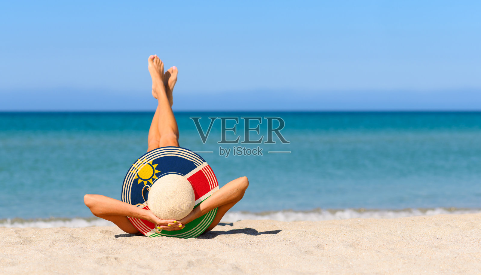 沙滩上一个苗条的女孩，戴着一顶代表纳米比亚国旗颜色的草帽。纳米比亚度假胜地的完美度假概念。照片摄影图片