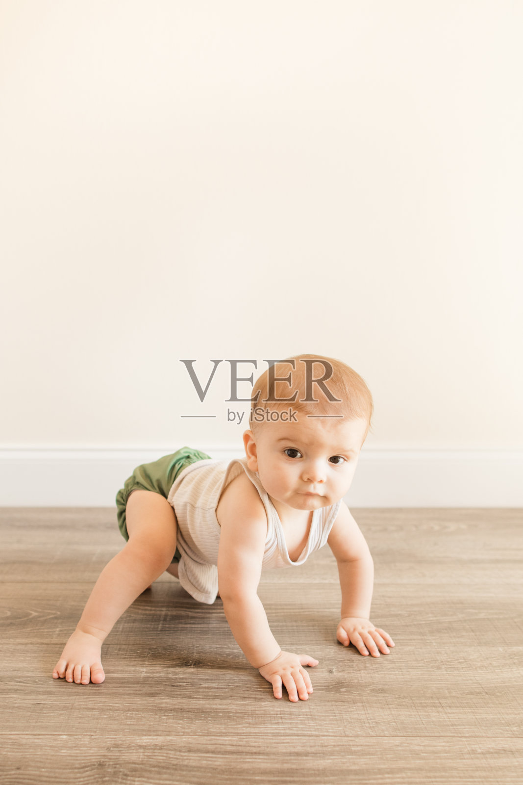 一名7个月大的患有多指畸形的大男婴，穿着橄榄绿可重复使用和可持续的布料尿布和米色背心，正在家里的木地板上学习爬行照片摄影图片