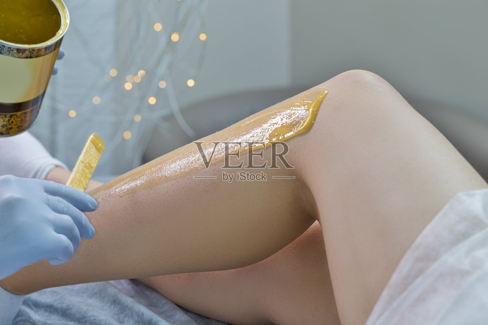 水疗中心的概念。美容师准备用刮刀在女性腿部涂抹液体蜡脱毛的特写。照片摄影图片
