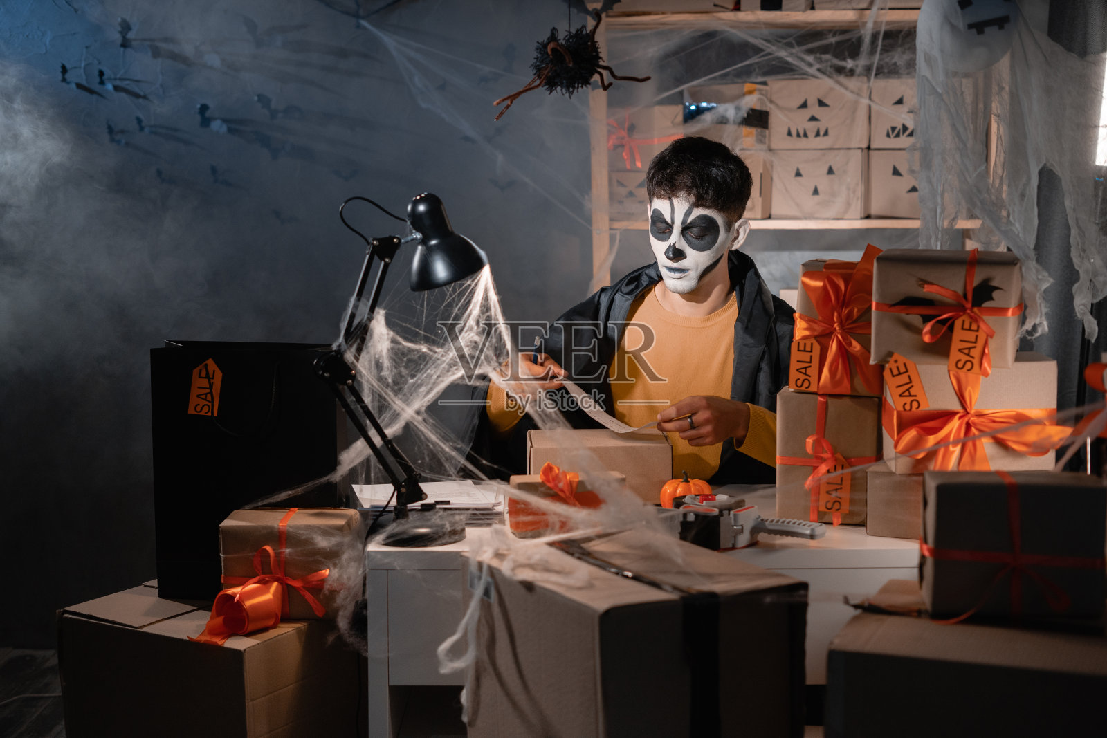 男妆骨架小企业主坐在一间装饰着祭奠亡灵的家仓库里。胶水的形式与盒子上的地址。万圣节销售和下降航运概念。副本的空间。照片摄影图片