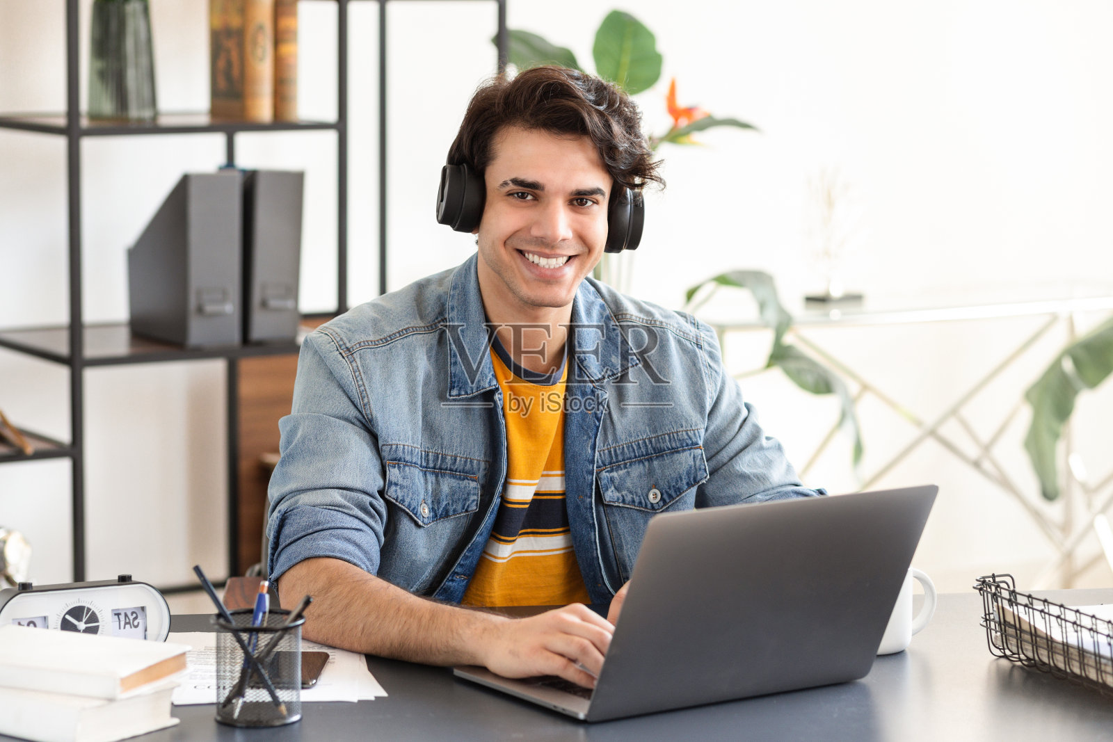 肖像快乐的男学生戴着耳机使用笔记本电脑，学习，远程学习，在线教育。年轻的自由职业者坐在家里的桌子旁，看着镜头工作，微笑着照片摄影图片