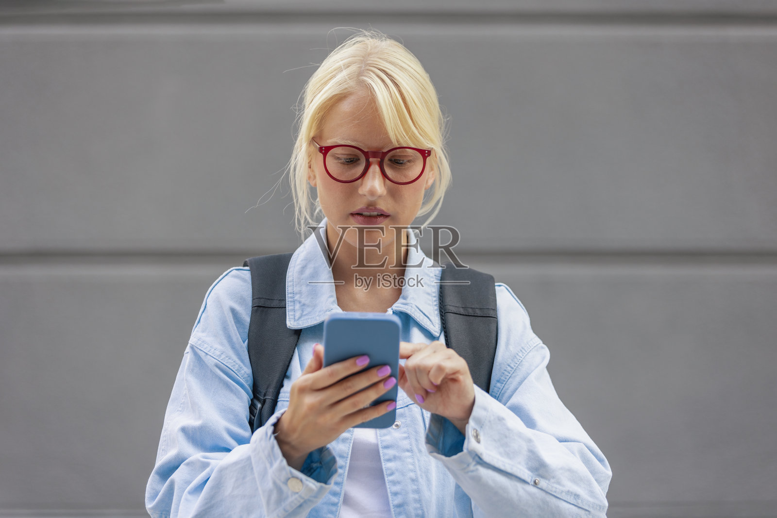 一个严肃的金发女学生手里拿着智能手机在屏幕上滚动，她身后是灰色的背景。照片摄影图片