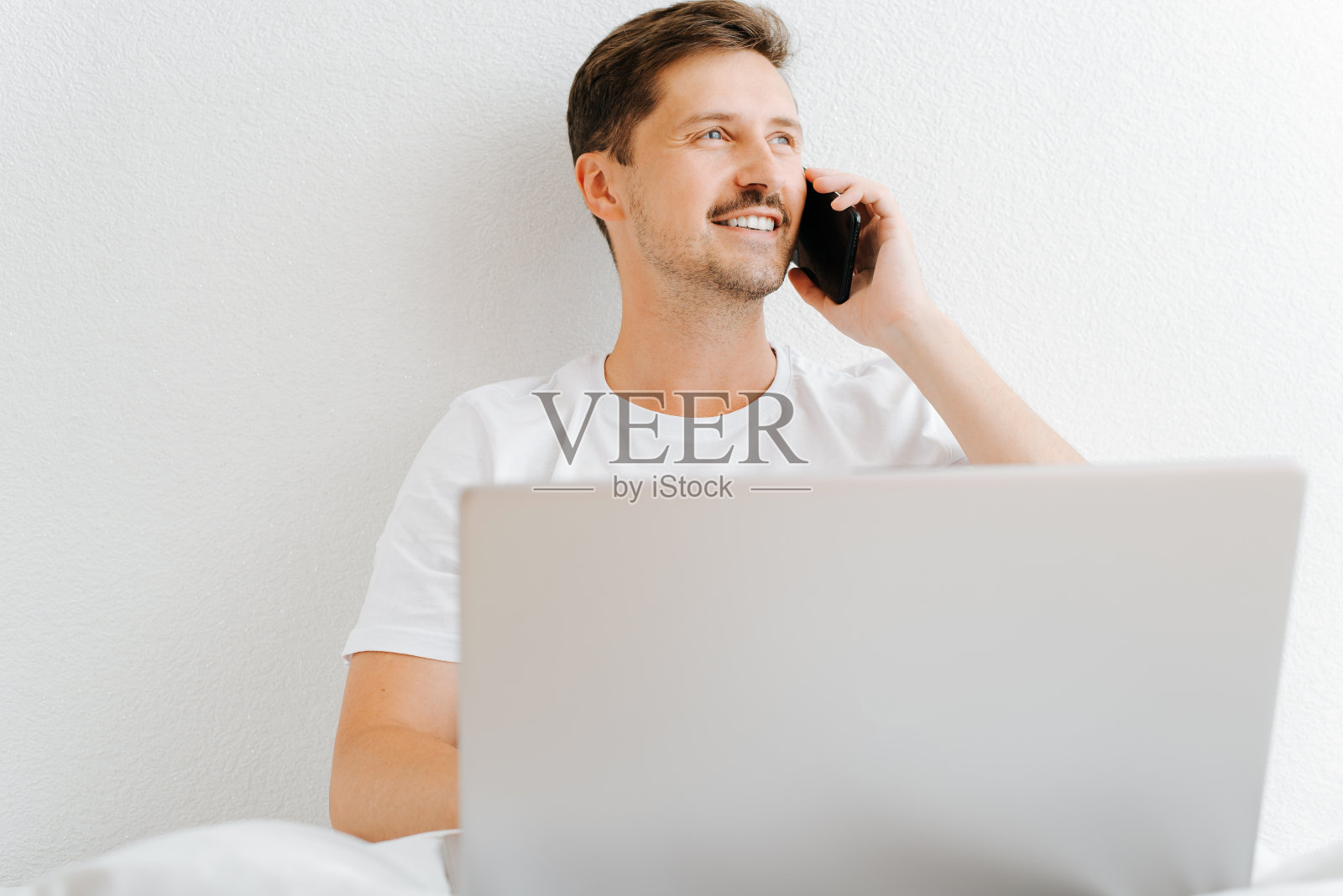 快乐英俊的年轻人在笔记本电脑上工作，在清晨坐在床上用智能手机打电话。微笑的男人在卧室里一边看别处一边用手机。在家远程工作照片摄影图片