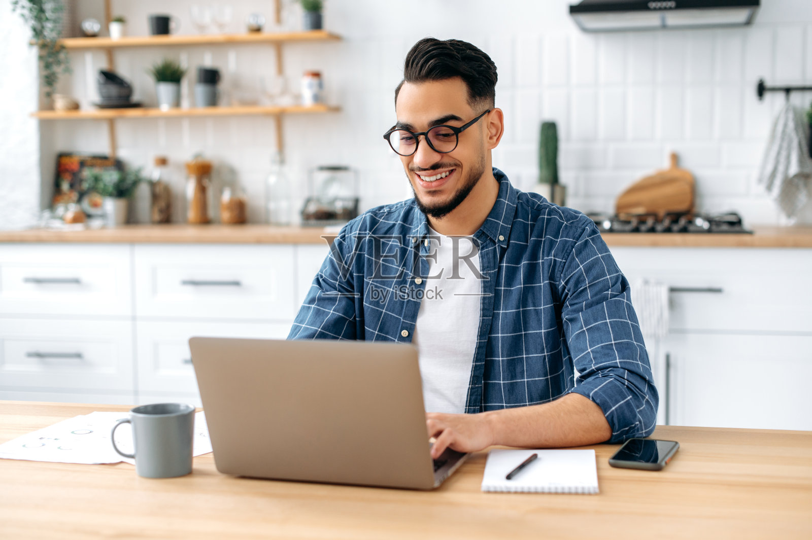 英俊积极的印度或阿拉伯男性企业家，自由职业者，远程工作时坐在家里的厨房，使用笔记本电脑，在键盘上打字，开发创造性的成功项目，微笑照片摄影图片